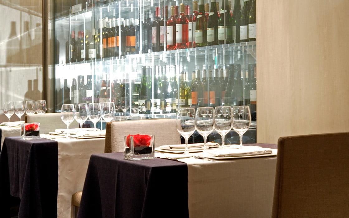 La fotografía muestra 3 mesas de un restaurante con copas encima. Detrás se puede ver una vitrina llena de botellas de cava y vino.